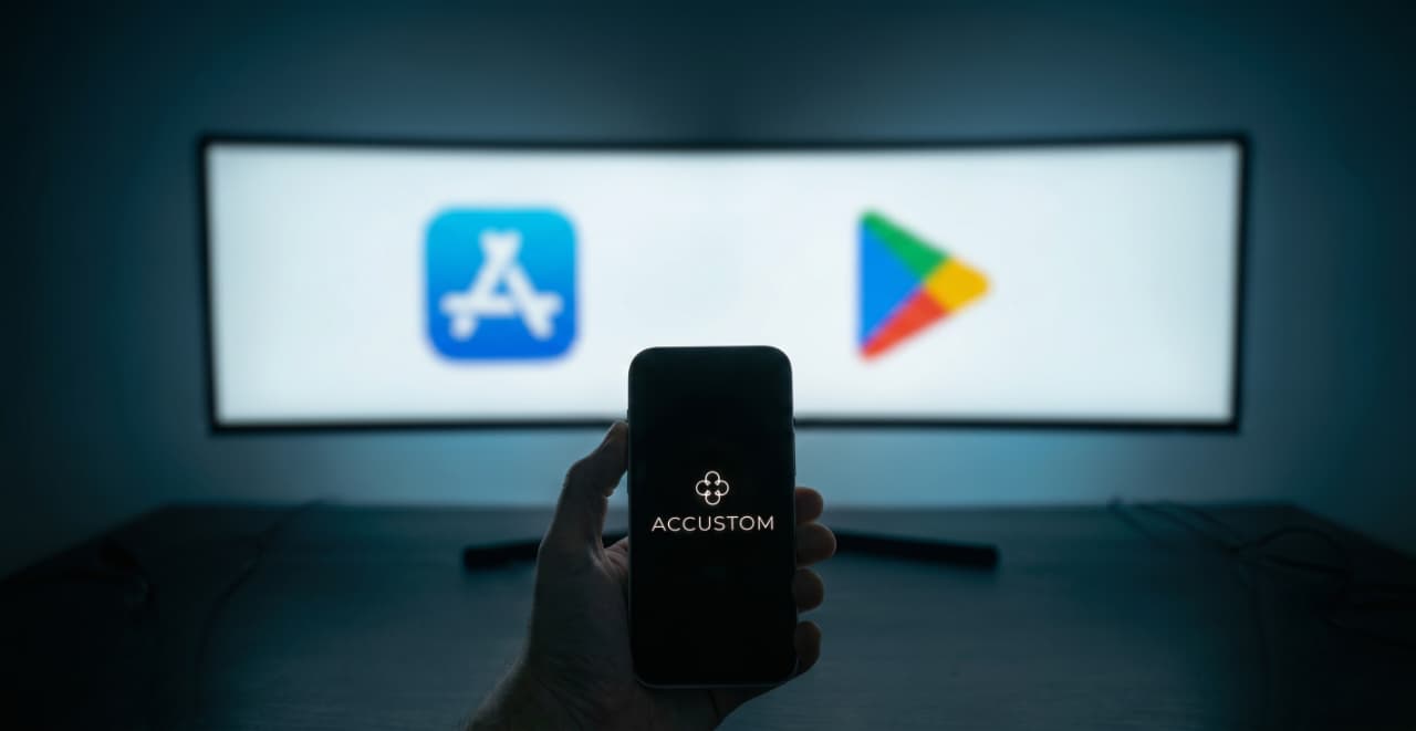 An Accustom app icon on a phone screen held up in front of a blurred Apple App Store and Google Play Store display